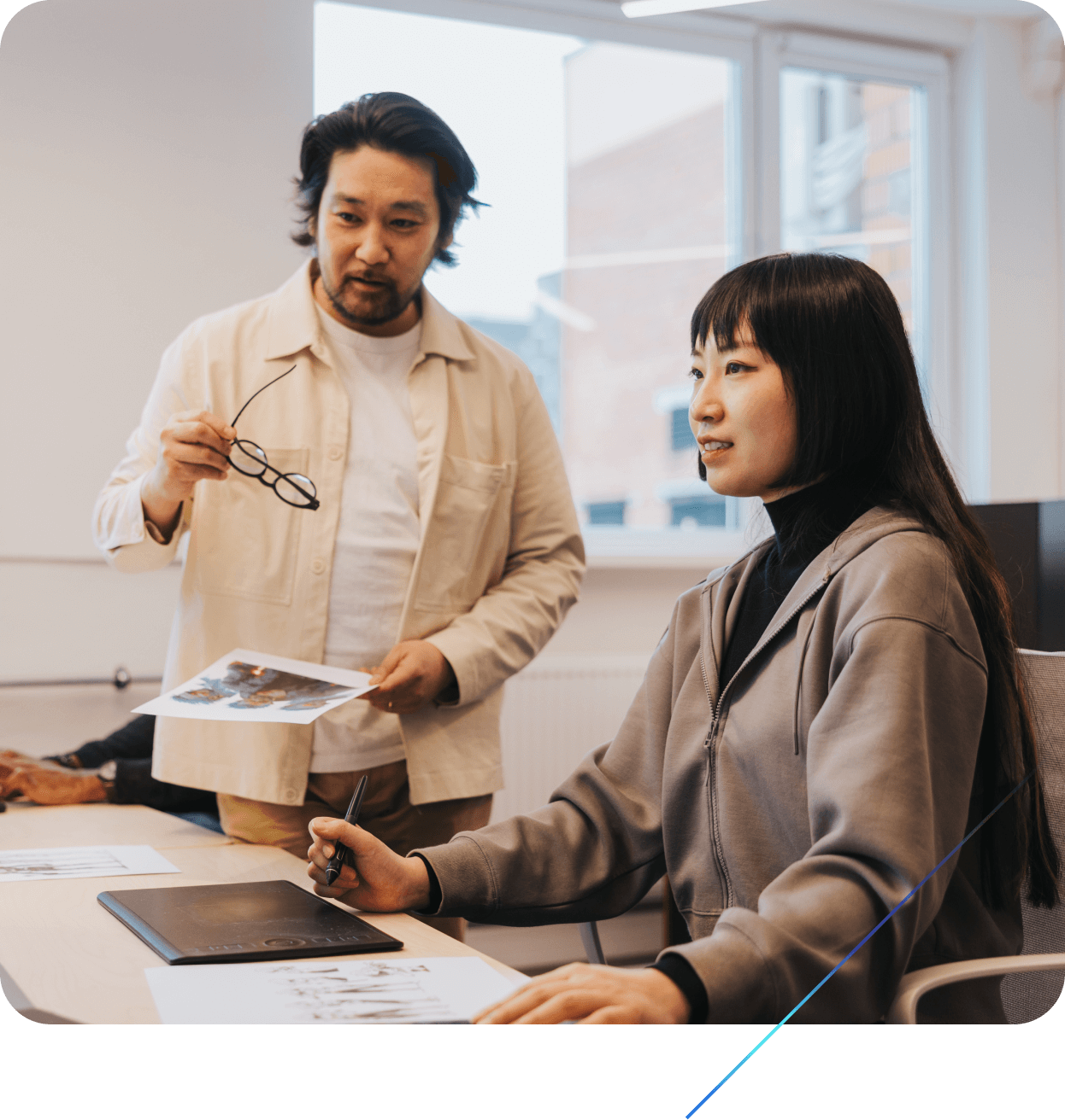 Two people at a desk discussing work with papers and a drawing tablet in an office setting.