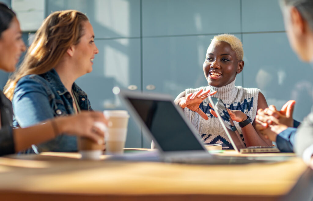Group of colleagues discussing work around a table with laptops in a bright office space.