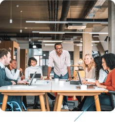 A diverse group of eight professionals is gathered around a long white conference table in a modern, open-plan office. 