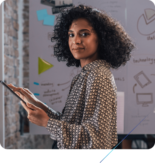 A woman with dark curly hair is shown in a professional setting, looking directly at the camera while holding a tablet in her hands. Behind her is a whiteboard with some diagrams and text.